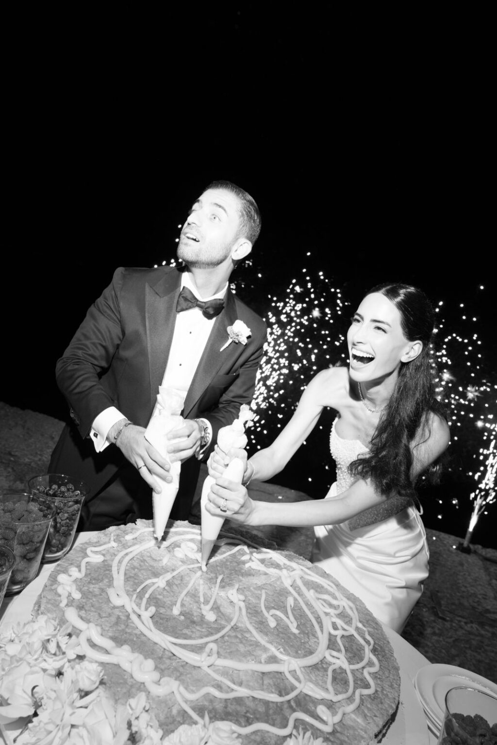 Eine Braut und ein Bräutigam dekorieren gemeinsam eine große Torte, während im Hintergrund ein Feuerwerk zu sehen ist.