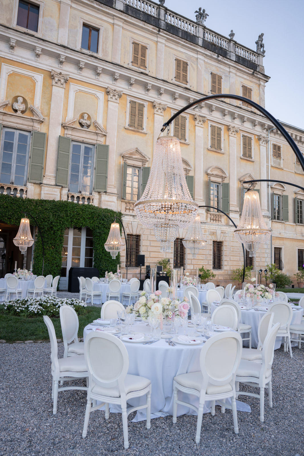 Elegante Hochzeitsfeier im Freien mit runden Tischen, weißen Stühlen und Kronleuchtern vor einer großen Villa.