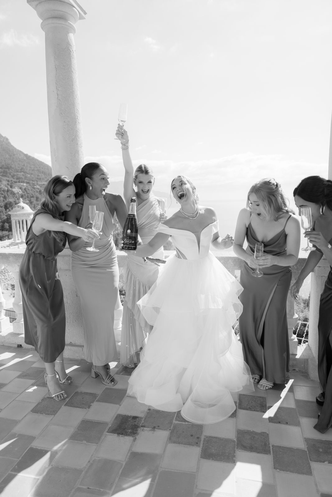 Eine fröhliche Braut und Brautjungfern lachen und feiern mit Champagner auf einer sonnigen Terrasse.