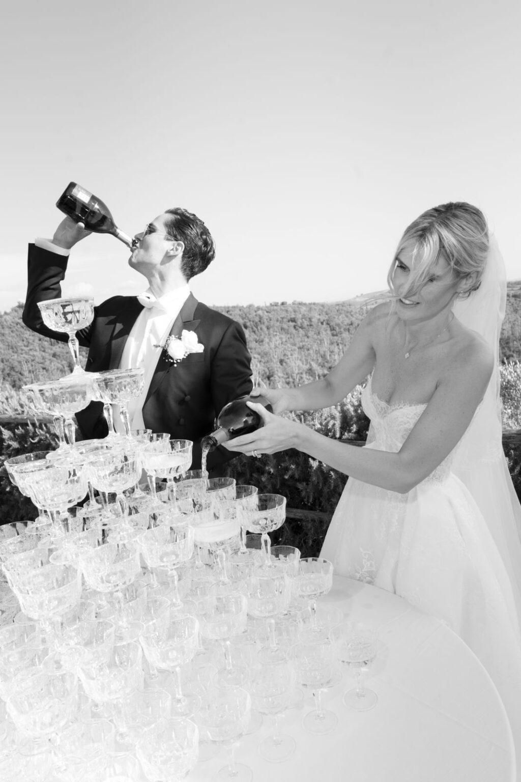 Braut und Bräutigam gießen Champagner in einen Glasturm im Freien; der Bräutigam trinkt aus der Flasche.