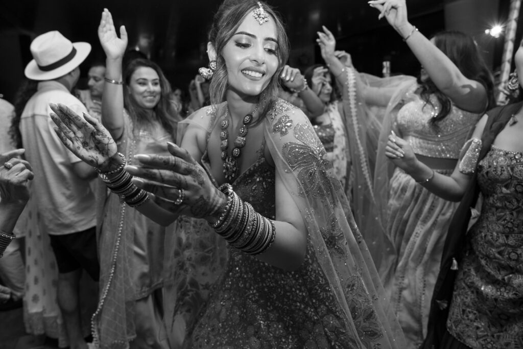 Eine Frau in traditioneller indischer Kleidung tanzt freudig auf einer Feier, umgeben von anderen Tanzenden.